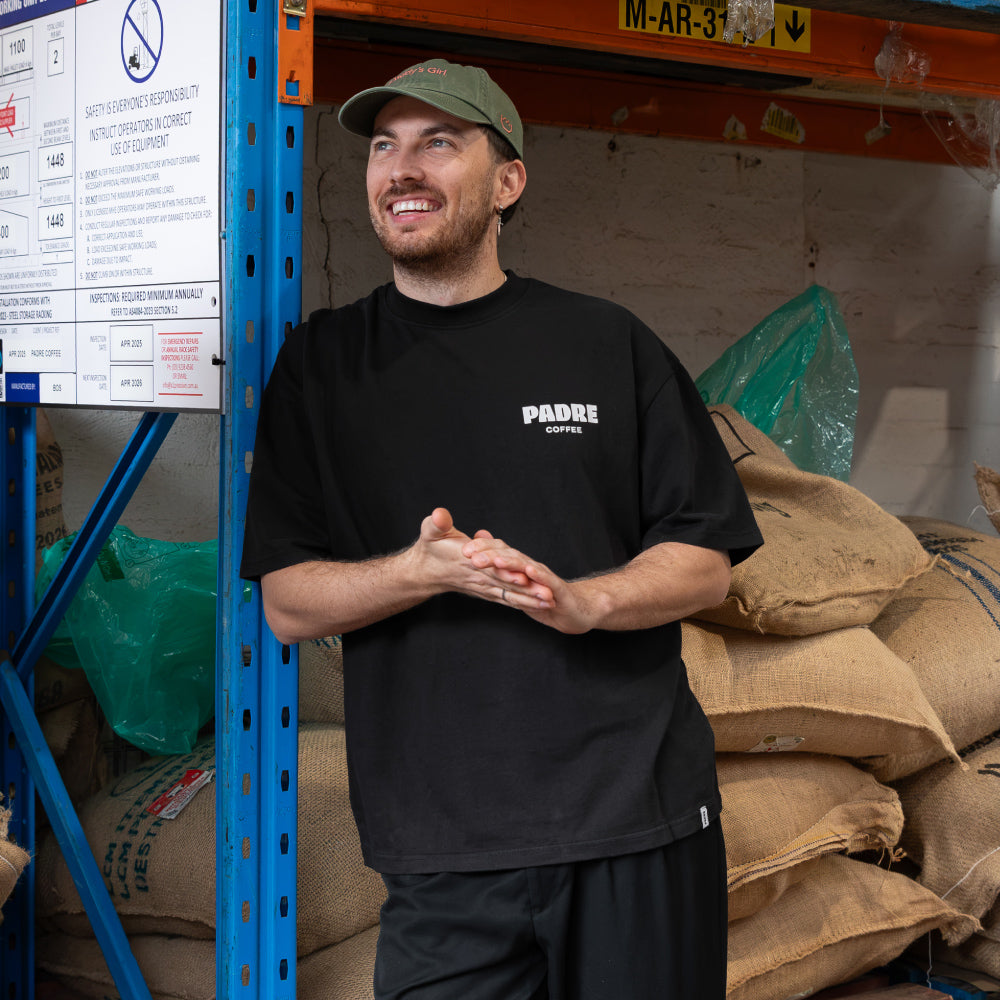 A man wearing a black t-shirt with 'PADRE COFFEE' printed on the chest and a green cap stands in a warehouse setting with coffee sacks on a shelf behind him.